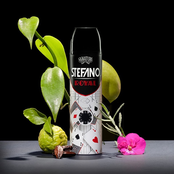 Un envase de desodorante en aerosol Stefano Royal decorado con motivos de cartas de póker, rodeado de grandes hojas verdes, una bergamota, dátiles y una flor rosa sobre un fondo negro.