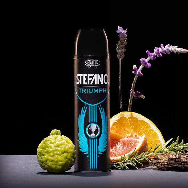 Un envase de desodorante en aerosol Stefano Triumph en el centro, acompañado de una bergamota, rodajas de naranja, una rama de romero y flores de lavanda sobre un fondo negro.