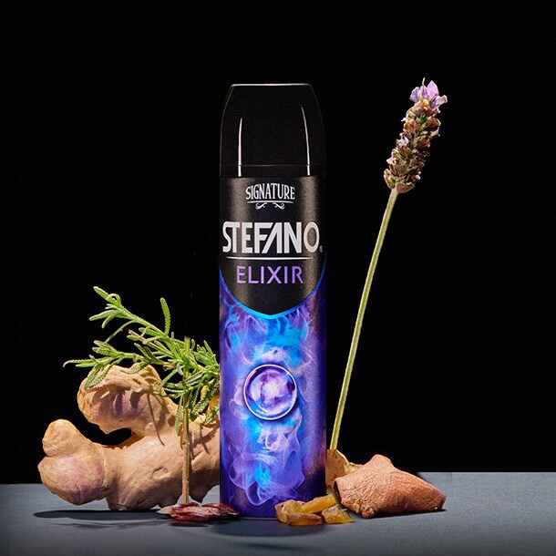 Un envase de desodorante en aerosol Stefano Elixir en el centro, acompañado de una raíz de jengibre, hojas verdes, trozos de resina y una flor de lavanda sobre un fondo negro.