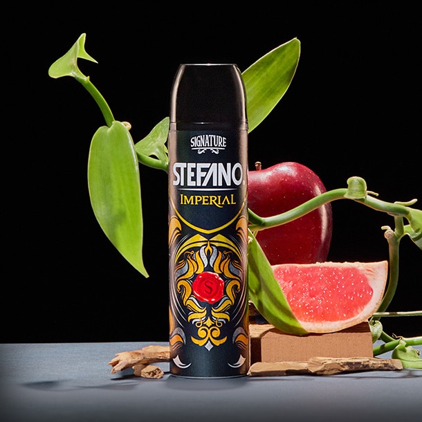 Un envase de desodorante en aerosol Stefano Imperial en el centro, rodeado de hojas de orquídea, una manzana roja, una rodaja de toronja y astillas de madera sobre un fondo negro.