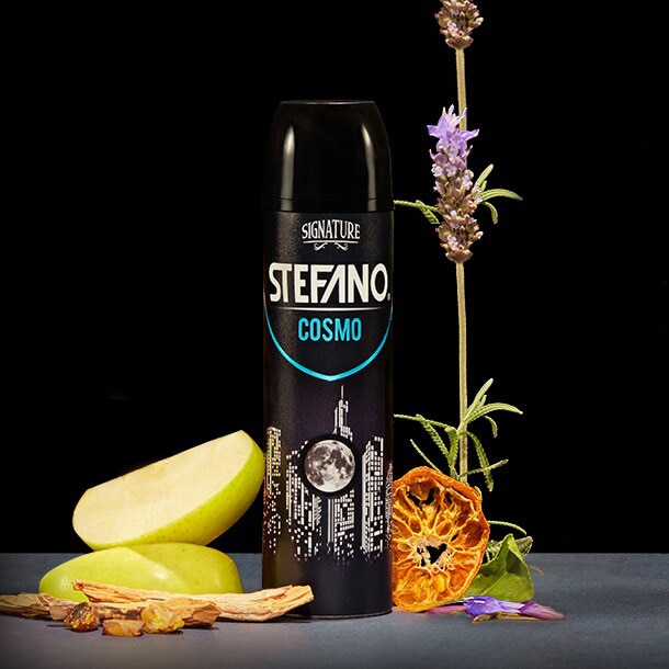 Un envase de desodorante en aerosol Stefano Cosmo en el centro, acompañado de rodajas de manzana verde, astillas de madera, una rodaja de naranja deshidratada y una flor de lavanda sobre un fondo negro.