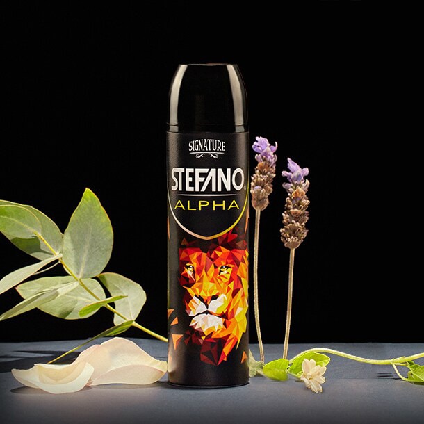 Un envase de desodorante en aerosol Stefano Alpha en el centro, decorado con la ilustración geométrica de un león y rodeado de hojas de eucalipto, pétalos claros y flores de lavanda sobre un fondo negro.